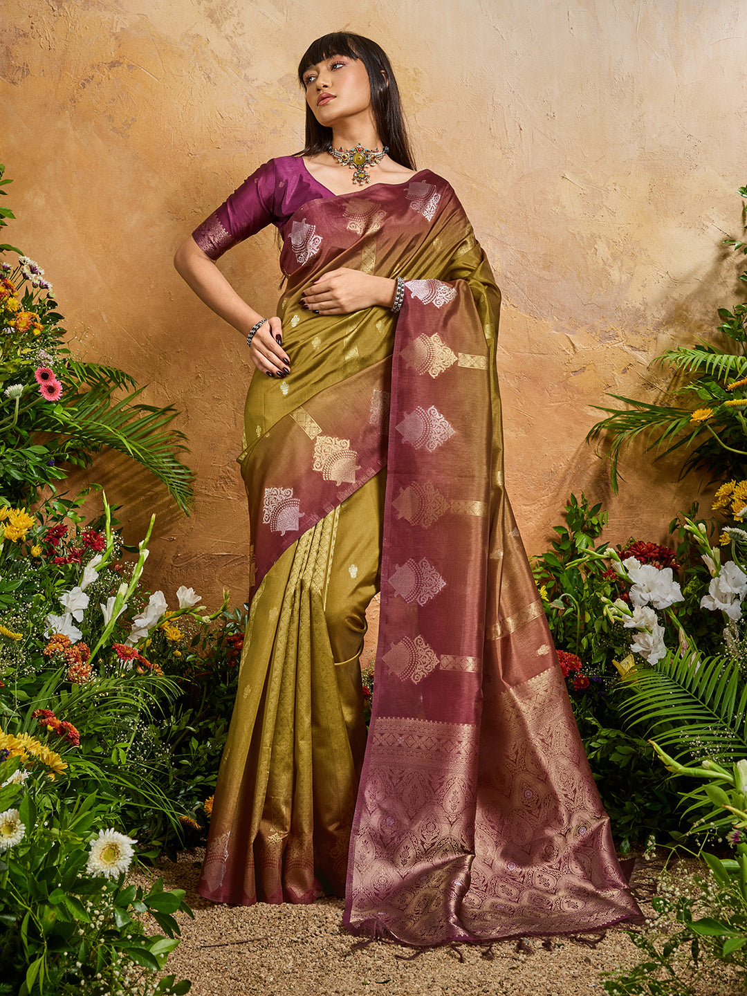 Woman in a green and purple saree standing among flowers with a beige background
