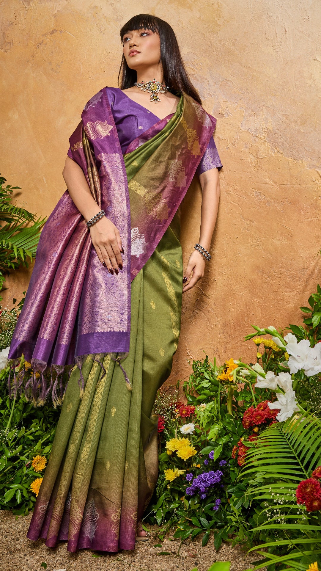 Woman in a green and purple saree standing among flowers and plants.