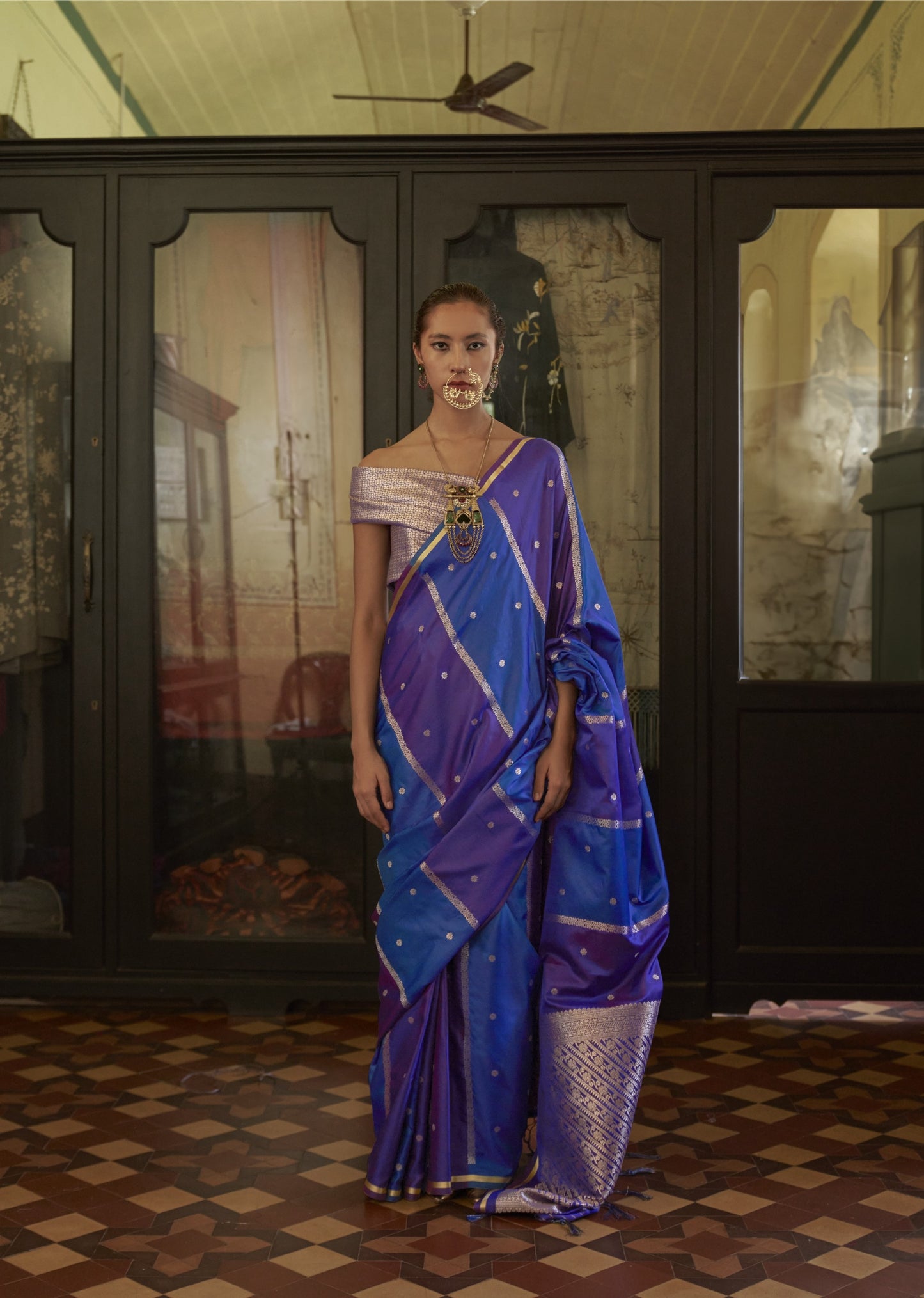 Woman in a blue saree standing in a room with a checkered floor and wooden wardrobe.
