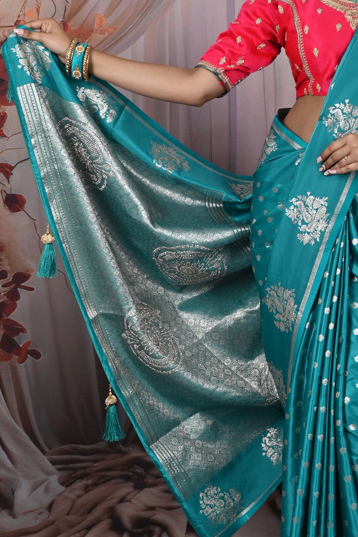 Woman in a red blouse and teal saree with white patterns against a draped curtain background