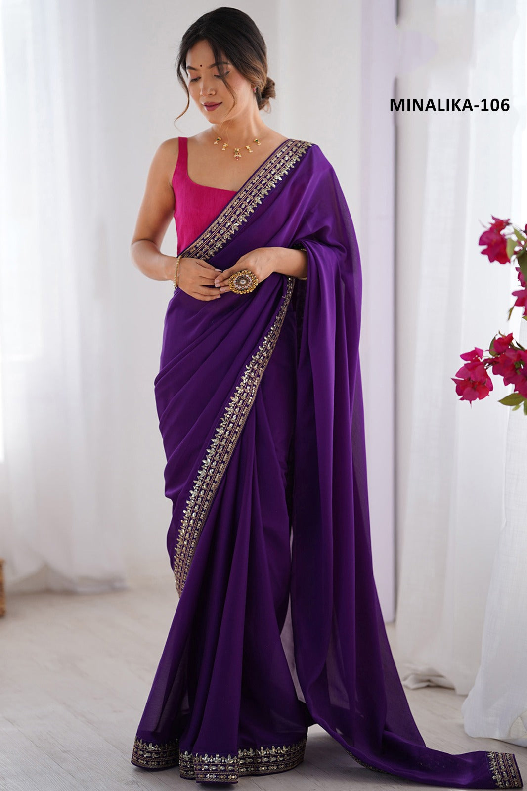Woman wearing a purple saree with gold border against a white curtain background