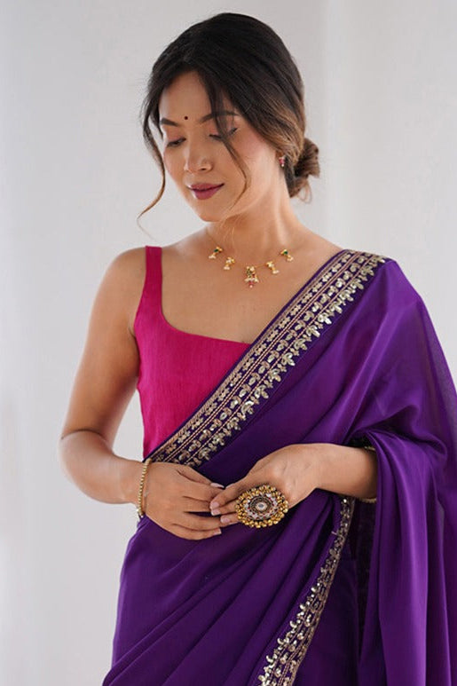 Woman wearing a purple saree with gold border against a white curtain background
