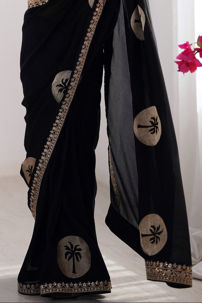 Woman wearing a black saree with white patterns, holding it in a room with white curtains and pink flowers.