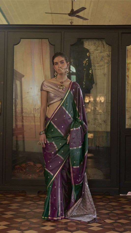 Woman in a traditional saree standing in a room with decorative walls and ceiling fan.